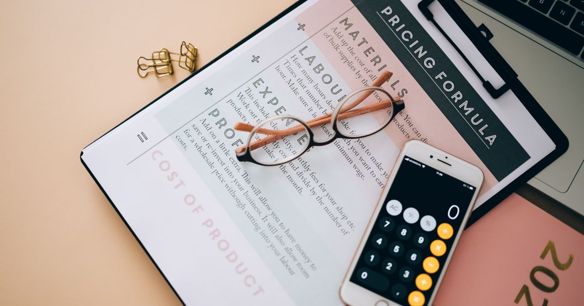 Overhead view of business tools including a phone calculator, pricing formula document, and eyeglasses on a desk.