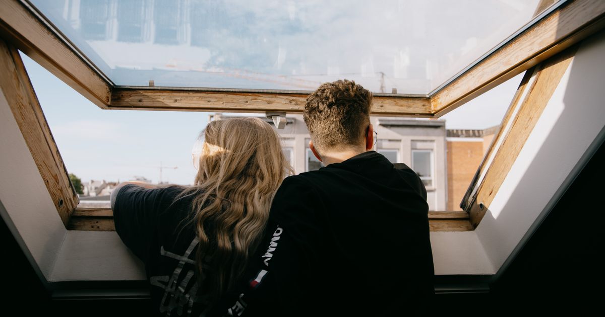 Back view of faceless couple in casual clothes hugging while standing near open window and looking at city