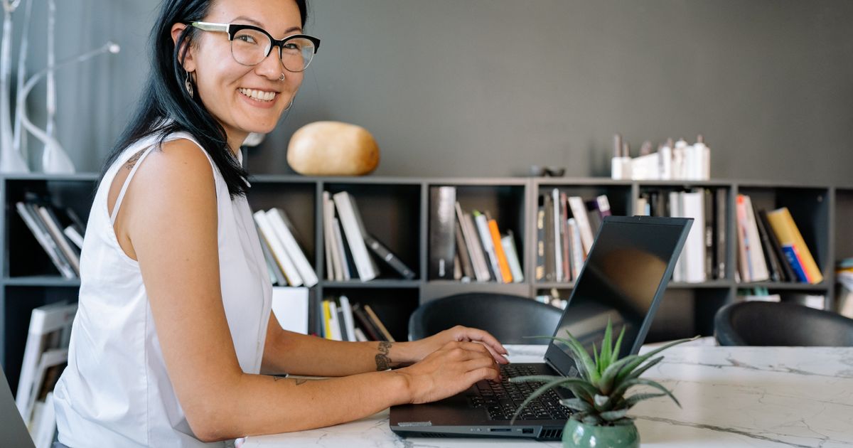 Cheerful businesswoman using laptop at a desk in a stylish office setup with bookshelves.