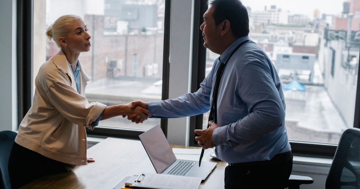 Young female candidate shaking hands with employer before job interview in light office with urban landscape through windows